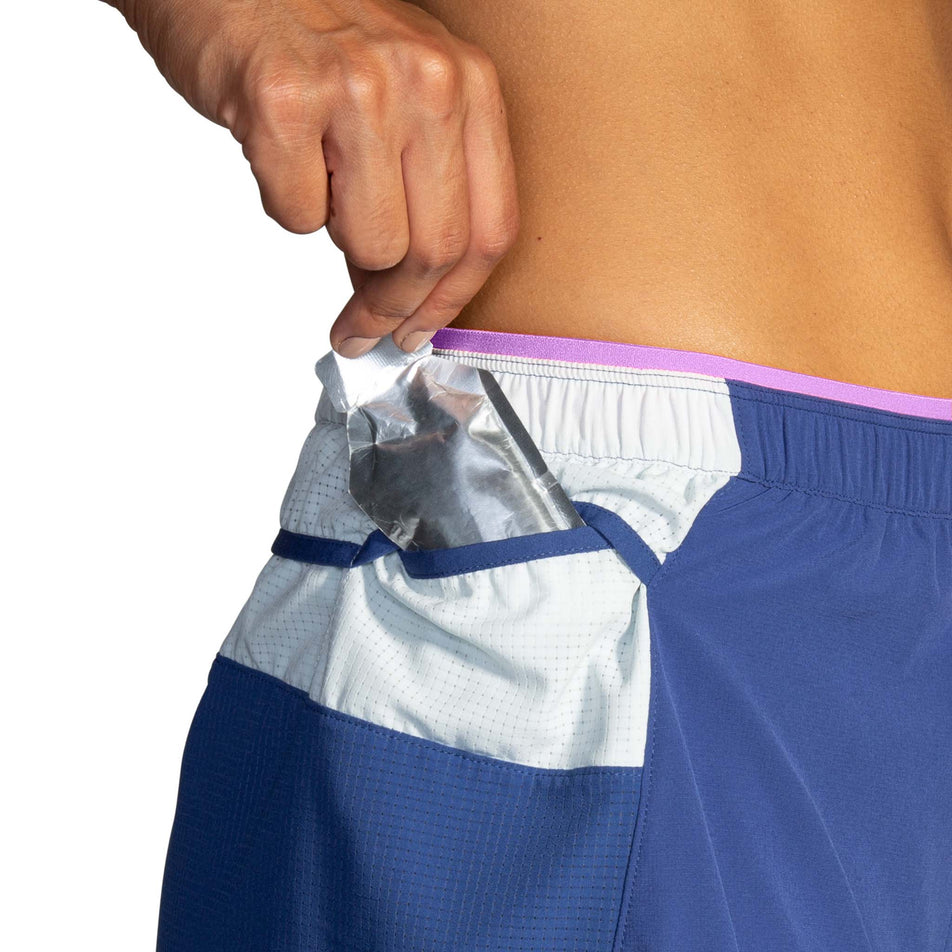 A model demonstrating that an energy gel can be stored in one of the mesh side pockets on the right-hip section of a pair of Brooks Women's High Point 3