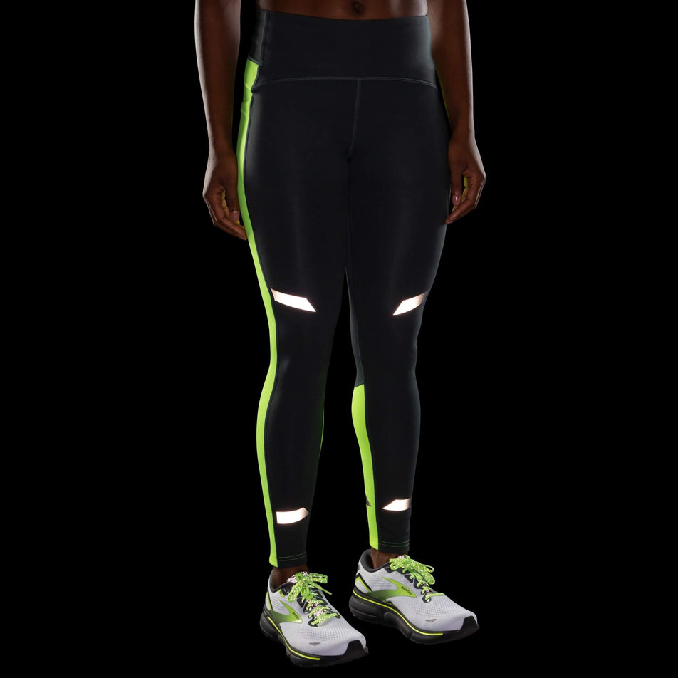 Front view of a model wearing a pair of Brooks Women's Run Visible Thermal Tights in the Asphalt/Nightlife colourway. Model is also wearing a pair of Brooks running shoes. Products are being worn in dark conditions to show the reflectivity. (8065569194146)
