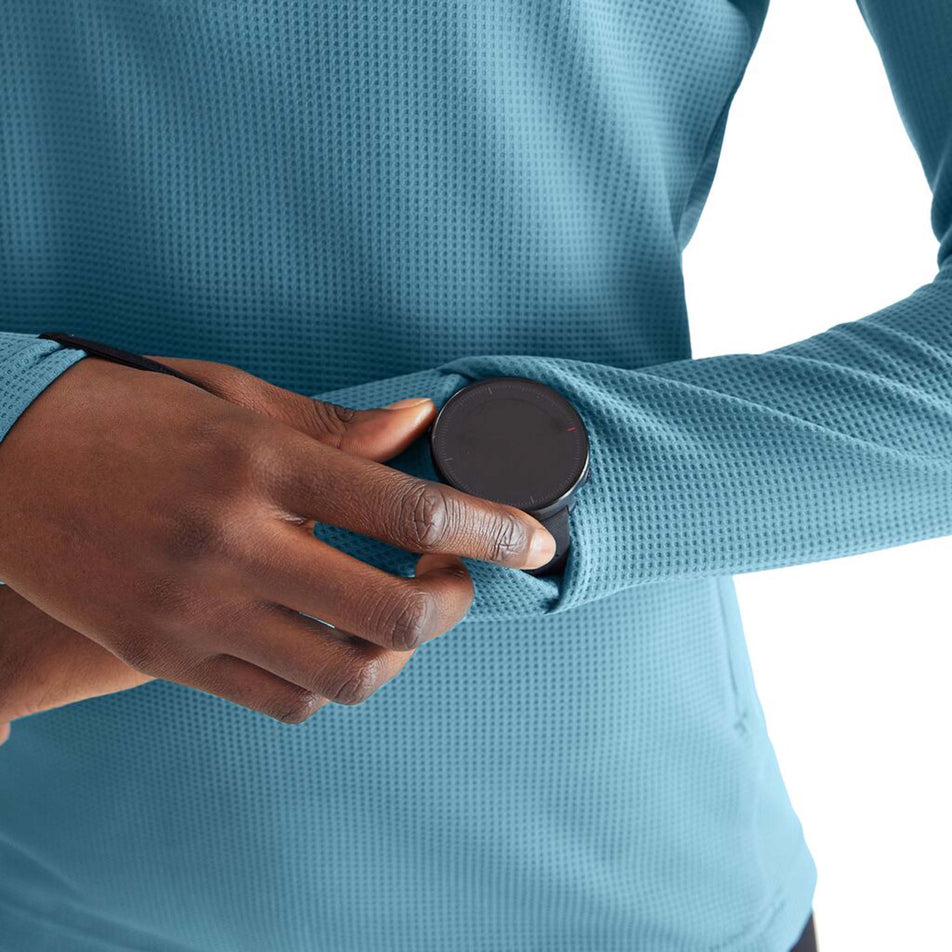 Close-up view of a model wearing an On Women's Climate Shirt in the Wash colourway, and demonstrating that a running watch fill fit through a 'watch window' in the cuff of the left sleeve.  (8004157046946)