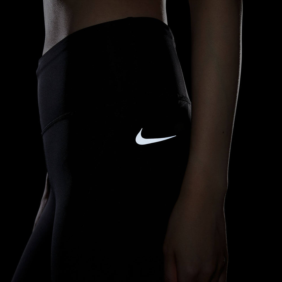 Left side view of the waist and hip section of a pair of Nike Women's Epic Fast Mid-Rise Pocket Running Leggings in the Black/Reflective Silv colourway. Leggings are being worn by a model in dark conditions to show the reflective Swoosh logo. (8049598562466)