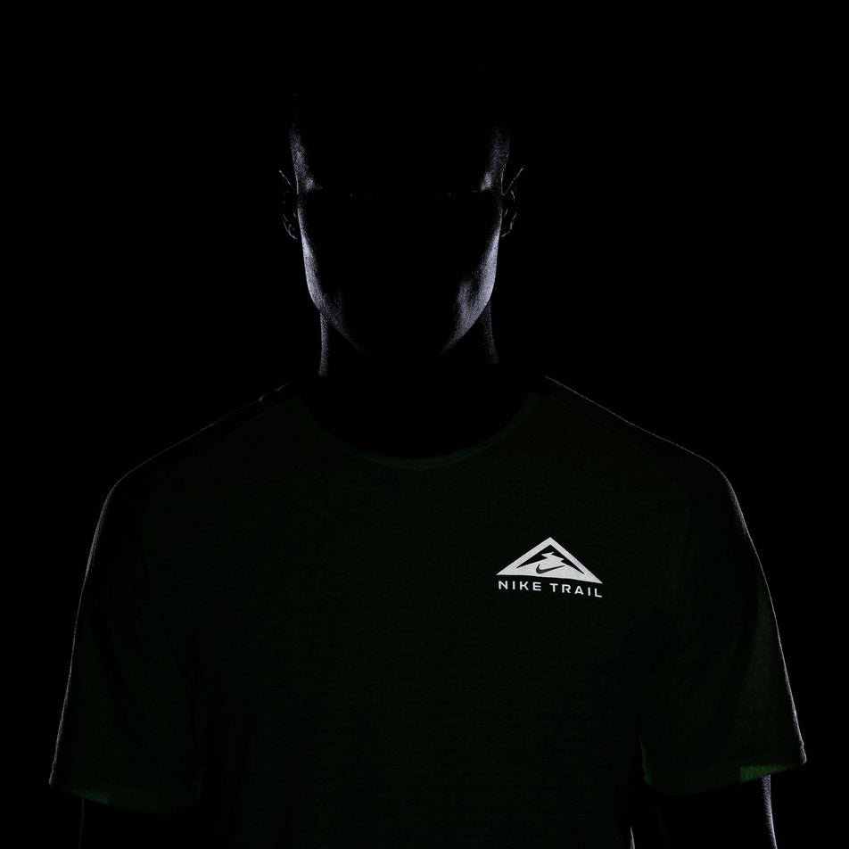 Close-up view of the reflectivity provided by the Nike Trail logo on the front of a Nike Men's Trail Solar Chase Dri-FIT Short-Sleeve Running Top in the Vapor Green/Black colourway.  (8215882825890)
