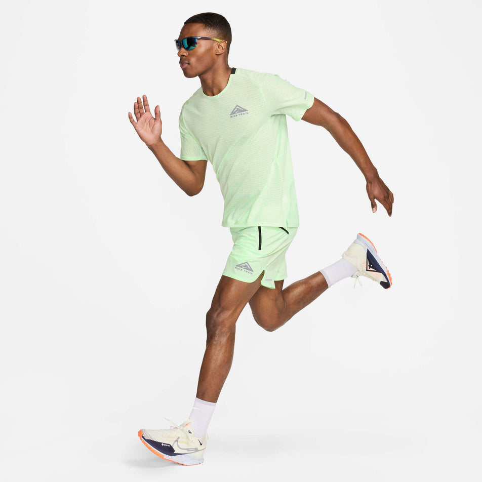 Front view of a model wearing a Nike Men's Trail Solar Chase Dri-FIT Short-Sleeve Running Top in the Vapor Green/Black colourway. Model is in a running motion and is also wearing Nike sunglasses, shorts, socks, and shoes. (8215882825890)