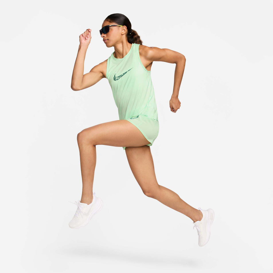 Side view of a model wearing a Nike Women's One Graphic Running Tank Top in the Vapor Green/Bicoastal colourway. Model is also wearing Nike sunglasses, shorts and shoes, and is in a running pose.  (8299396104354)