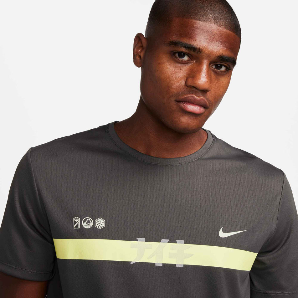Close-up front view of the upper third of a Nike Men's Miler Dri-FIT UV Short-Sleeve Running Top in the Medium Ash/Luminous Green/Light Bone colourway. Top is being worn by a model. (8141458473122)