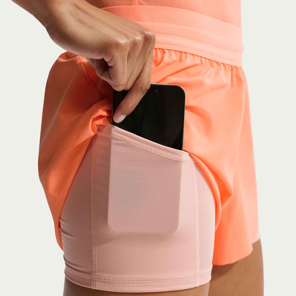 A model demonstrating the stash pocket on the side of the Nike Women's Swift Dri-FIT Mid-Rise 2-in-1 Running Shorts in the Orange Pulse/Reflective Silver colourway.