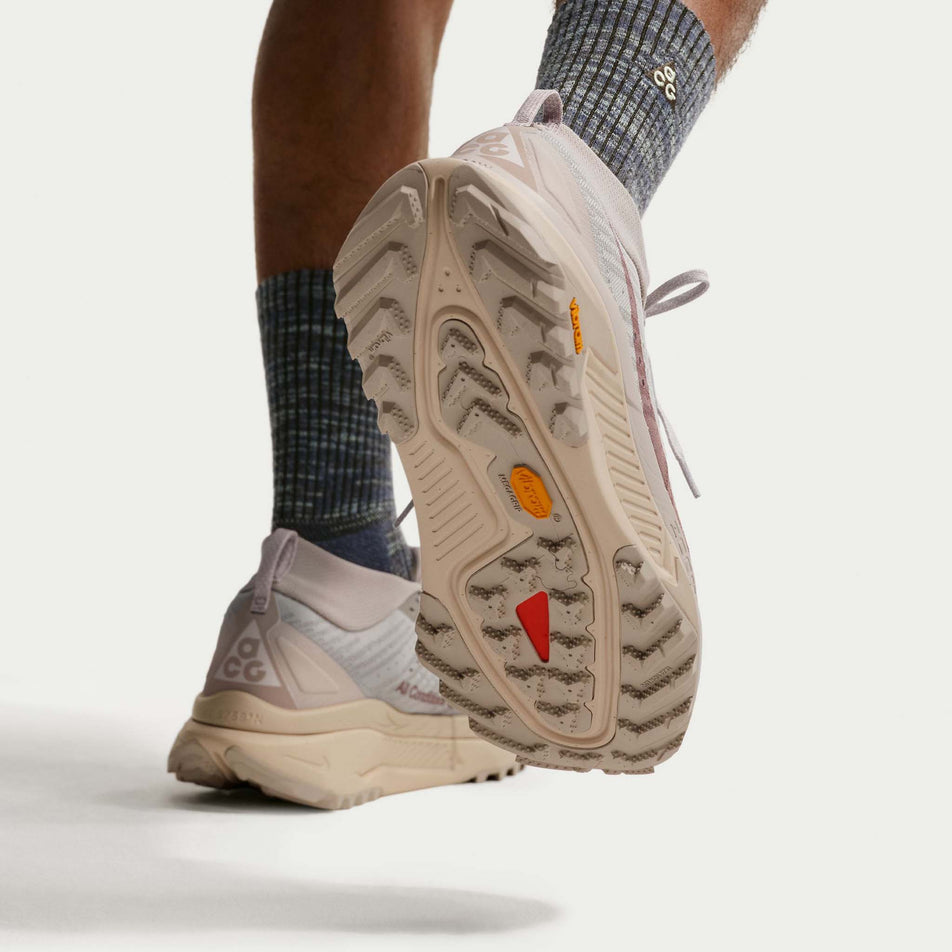A model wearing a pair of Nike Men's ACG Zegama Trail Running Shoes in the Cream II/Diffused Taupe-Light Orewood Brown colourway.