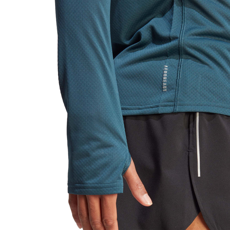 Side view of a model wearing an adidas Women's Run Icons Running Long-Sleeve Top in the Arctic Night colourway. Lower right side of the top is visible, showing the Aeroready call-out and the thumb loop in the sleeve.  (8005336432802)