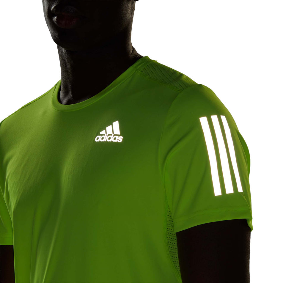Close-up front view of a model wearing an adidas Men's Own the Run Tee in the Lucid Lemon colourway. Upper half of the t-shirt is visible in low light conditions, so the reflective adidas logo on the chest, and the reflective three stripes on the sleeve are visible.  (8005317394594)