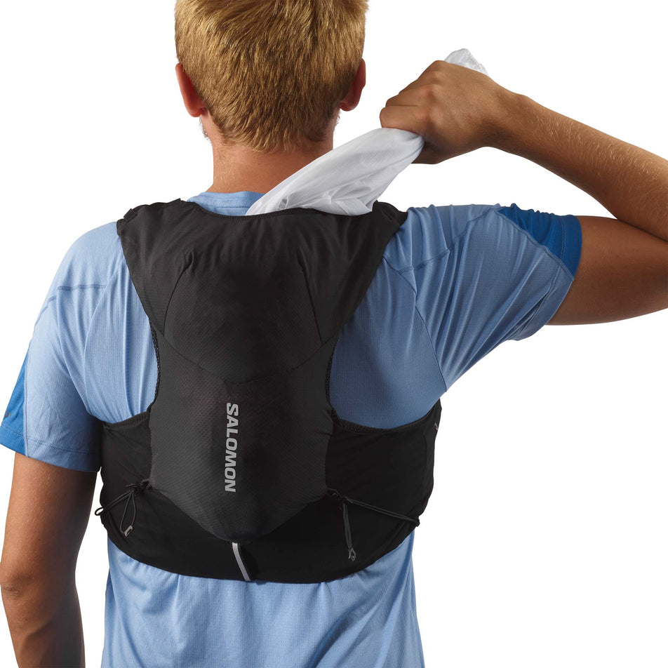 Back view of a model wearing and taking a top out of a Salomon Unisex ADV Skin 5 Running Vest in the Black/Ebony colourway, with flasks included. (7991863115938)