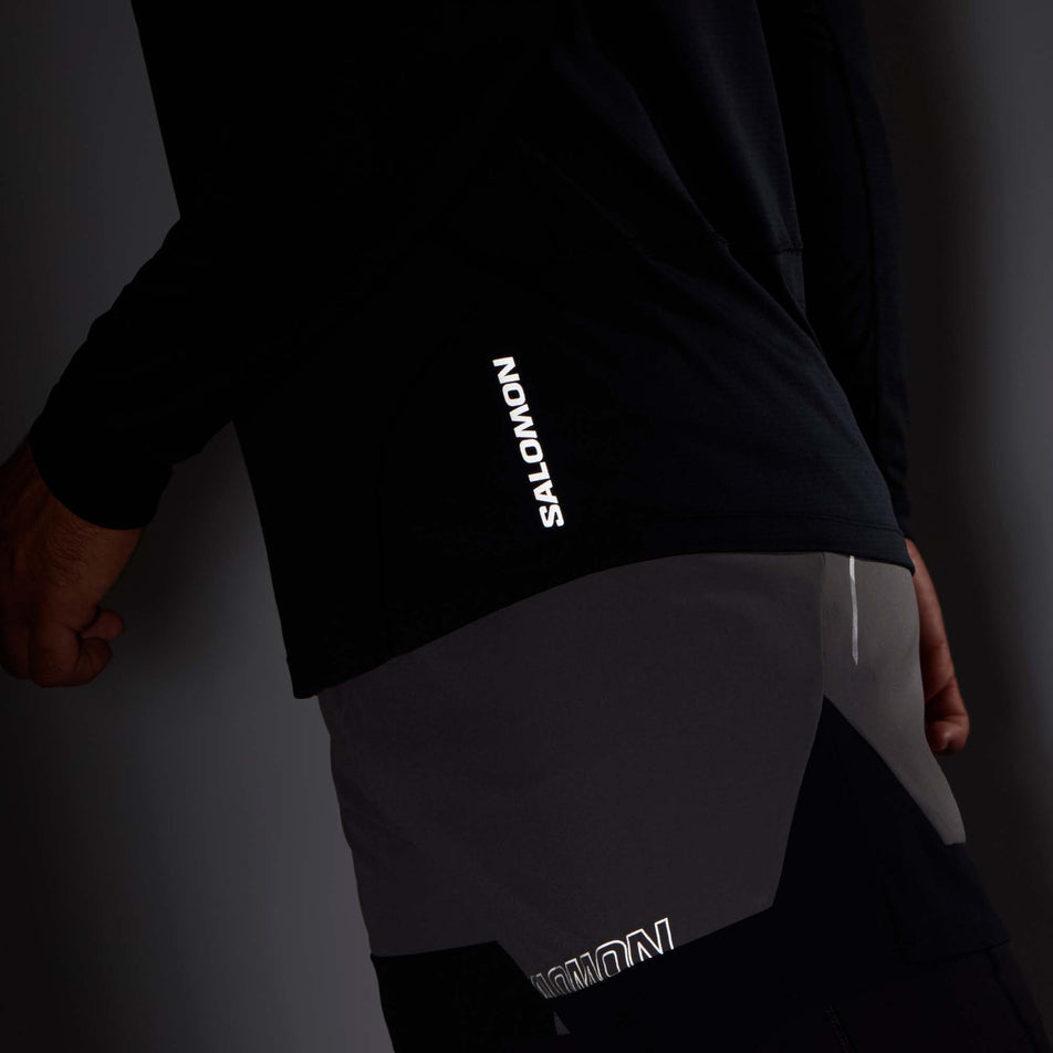 Close-up view of the lower left section of a Salomon Men's Cross Run Long Sleeve T-Shirt in the Deep Black colourway. Worn by a model in low light conditions to show the reflective Salomon logo on the fabric. (8008540324002)