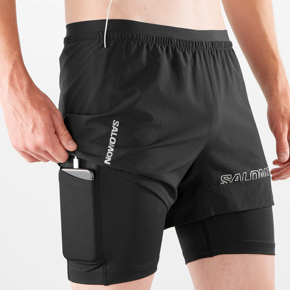 A model demonstrating that a phone can be stored in the pocket on the outside of the inner liner on the right leg. The cable to headphones can exit out of a hole near the waistband. The model is wearing a pair of Salomon Men's Cross 2in1 Shorts in the Deep Black colourway. (8008550121634)