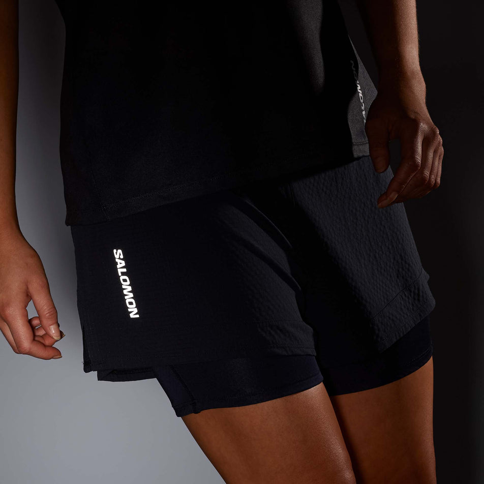 Front view of a model wearing a pair of Salomon Women's Sense 2in1 Shorts in the Deep Black colourway, in low light conditions to show the reflective Salomon logo. (8000776437922)