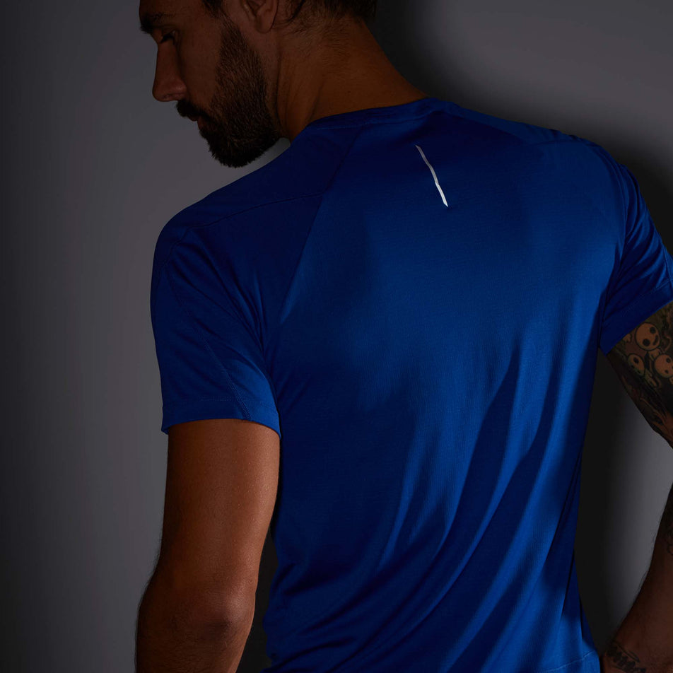 Close-up back view of a model wearing a Salomon Men's Cross Run Short Sleeve T-Shirt in the Surf The Web colourway. T-shirt is being worn in low light conditions to show the reflective strip in the middle of the upper back section.  (8008549040290)