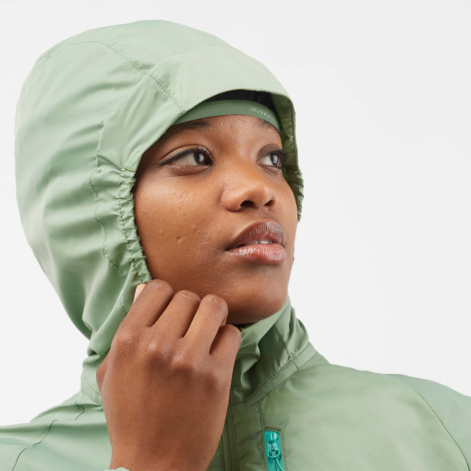 Close-up front view of a model wearing a Salomon Women's Bonatti Cross Wind Jacket in the Lily Pad/Deep Lichen Green colourway. The hood is up.  (7999055462562)