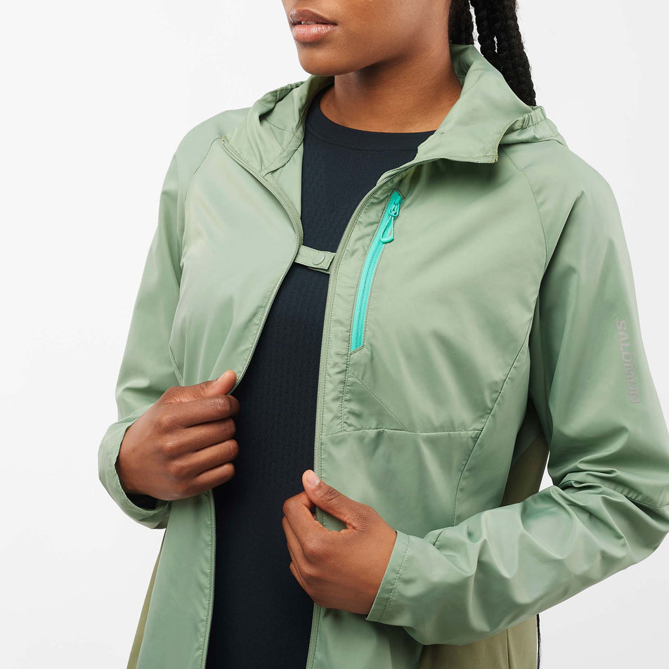 Close-up front view of a model wearing a Salomon Women's Bonatti Cross Wind Jacket in the Lily Pad/Deep Lichen Green colourway. Jacket is unzipped but held partially closed with the front clip. (7999055462562)