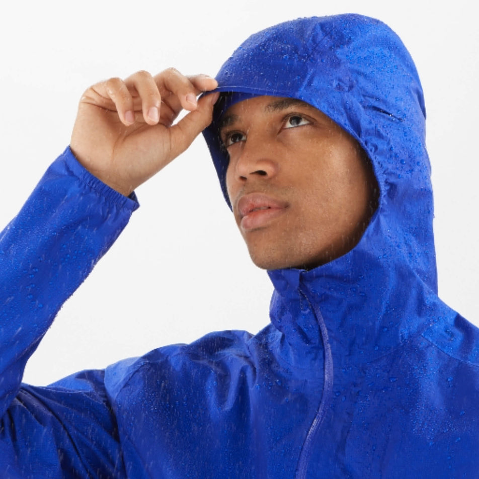 Close-up view of a model wearing a Salomon Men's Bonatti Waterproof Jacket in the Surf The Web colourway. Upper part of the jacket can be seen in the image. Model is wearing jacket with the hood up. Beads of water are sitting on the fabric. (8008537833634)
