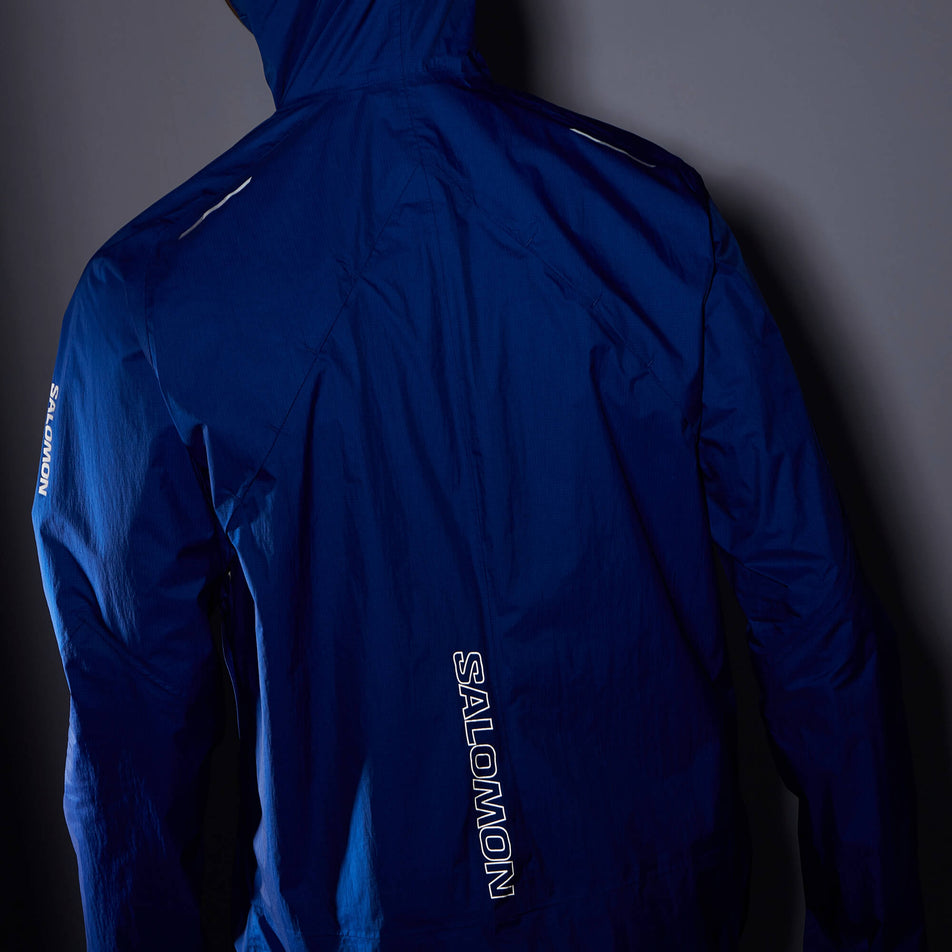 Back view of a model wearing a Salomon Men's Bonatti Waterproof Jacket in the Surf The Web colourway. The image was taken in low light conditions to show the reflective Salomon logo in the lower centre part of the back, and the reflective strip on the left and right shoulder. (8008537833634)