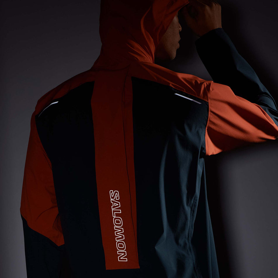 Close-up back view of a model wearing a Salomon Men's Bonatti Trail Jacket in the Burnt Ochre/Carbon colourway. Jacket is being worn with the hood up in low light conditions to show the reflective Salomon logo in the middle of the back, and the two reflective strips on the upper left and right parts of the back. (8008561098914)
