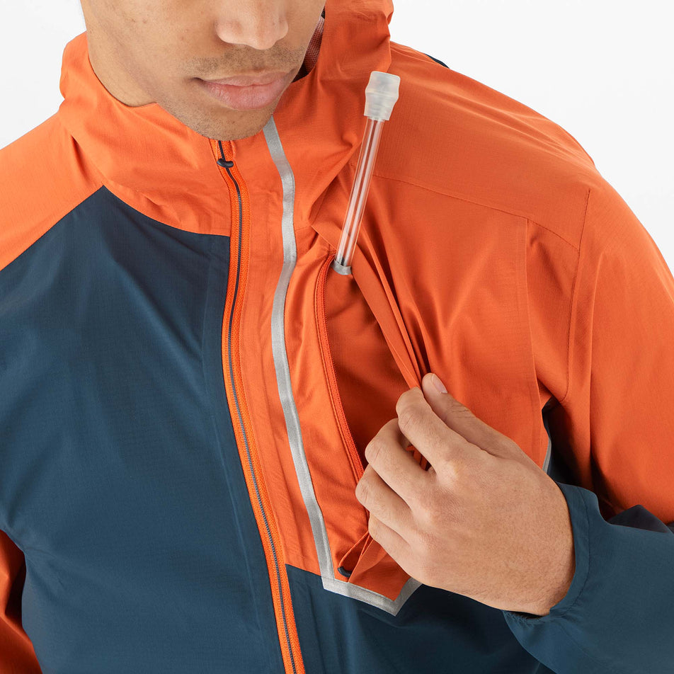 Close-up front view of a model wearing a Salomon Men's Bonatti Trail Jacket in the Burnt Ochre/Carbon colourway. Model is demonstrating that the tube from a drinks flask can be fed through the inner liner of the chest pocket - allowing the valve to be accessible when the jacket is worn over the top of a hydration vest.  (8008561098914)