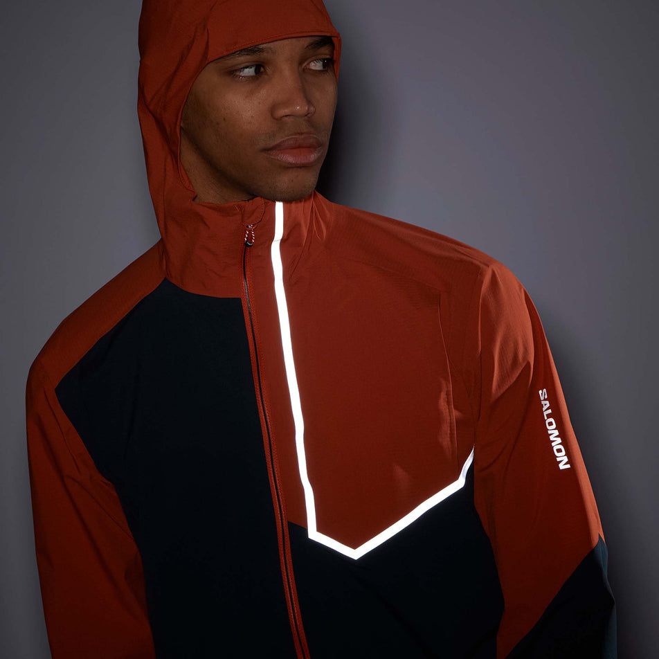 Close-up front view of a model wearing a Salomon Men's Bonatti Trail Jacket in the Burnt Ochre/Carbon colourway. Jacket is being worn with the hood up in low light conditions to show the reflectivity on the left side of the chest and the left sleeve. (8008561098914)