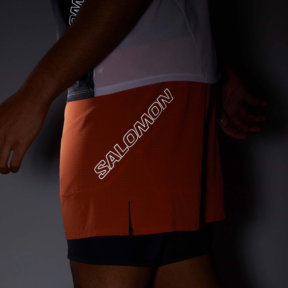 Close-up side view of a model wearing a pair of Salomon Men's Sense Aero 2in1 Shorts in the Burnt Ochre colourway. Being worn in low light conditions to show the reflective Salomon logo on the left-side of the shorts. (8008583315618)