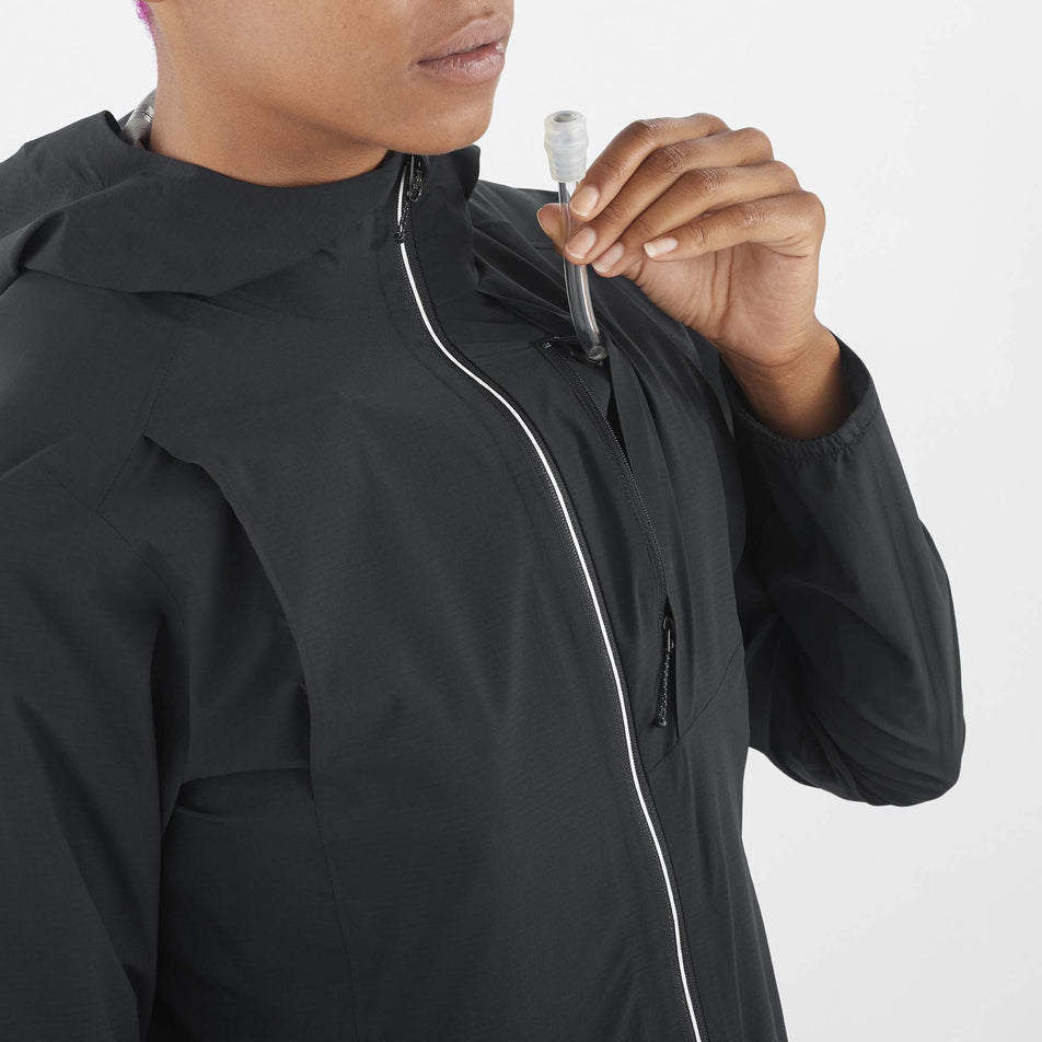 Close-up front view of a model wearing a Salomon Women's Bonatti Trail Jacket in the Deep Black colourway. Model is demonstrating that the tube from a soft flask can come through the front zipped chest pocket. (8157861019810)