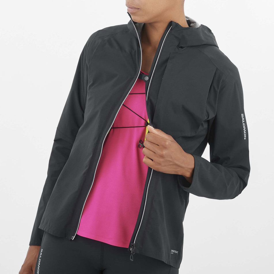 Close-up front view of a model wearing a Salomon Women's Bonatti Trail Jacket in the Deep Black colourway. Model has the jacket unzipped but the front sides are attached to each other via the fastener at the chest.  (8157861019810)