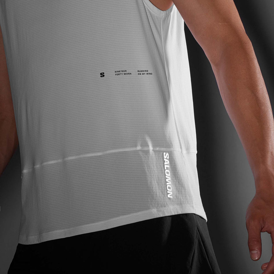 Close-up front view of the logo and brand callout  on the front left side of a Salomon Men's Cross Run Tank GFX in the White colourway. Top is being worn by a model in low light conditions. (8246399729826)