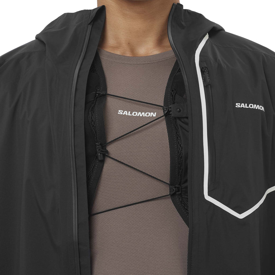 Front view of a model wearing the Salomon Men's Bonatti Pro Shell Jacket in the Deep Black colourway with a hydration vest underneath. 
