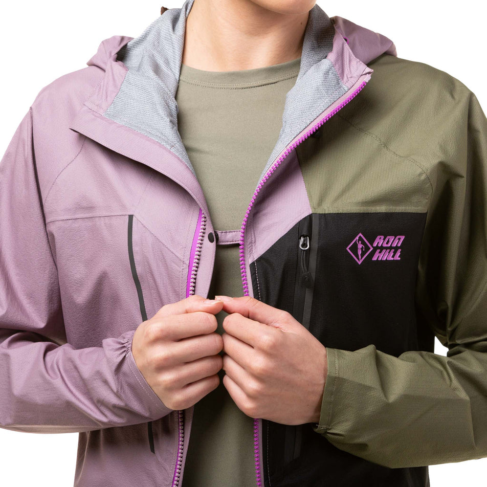 Close-up front view of a model demonstrating that, on a Ronhill Women's Tech Fortify Jacket, the front left and right sides can be fastened together with a built-in clip - when the jacket is unzipped. Jacket is in the Woodland/Stardust/Black colourway. (8024348328098)