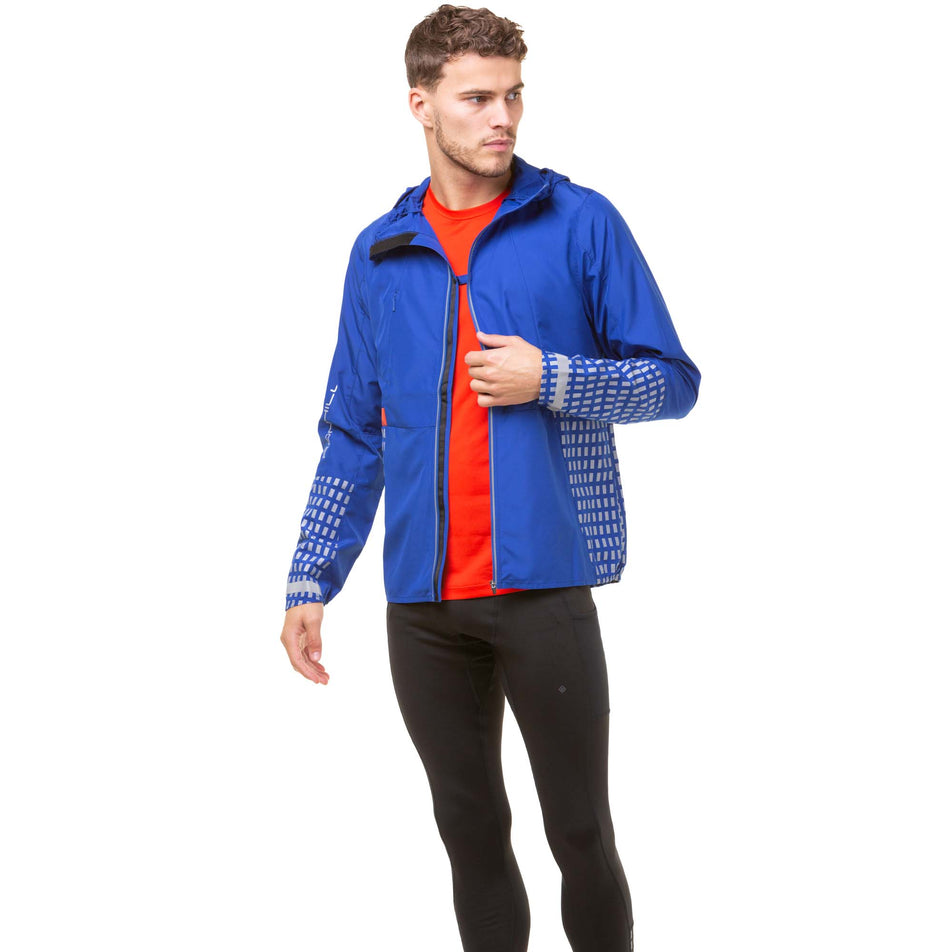 Front view of a model wearing a Ronhill Men's Tech Afterhours Jacket in the Cobalt/Flame/Reflect colourway. Jacket is unzipped, with the left and right front sides clipped together with a fastener at the chest. Model also wearing a red Ronhill running top and black Ronhill tights.  (8032156385442)