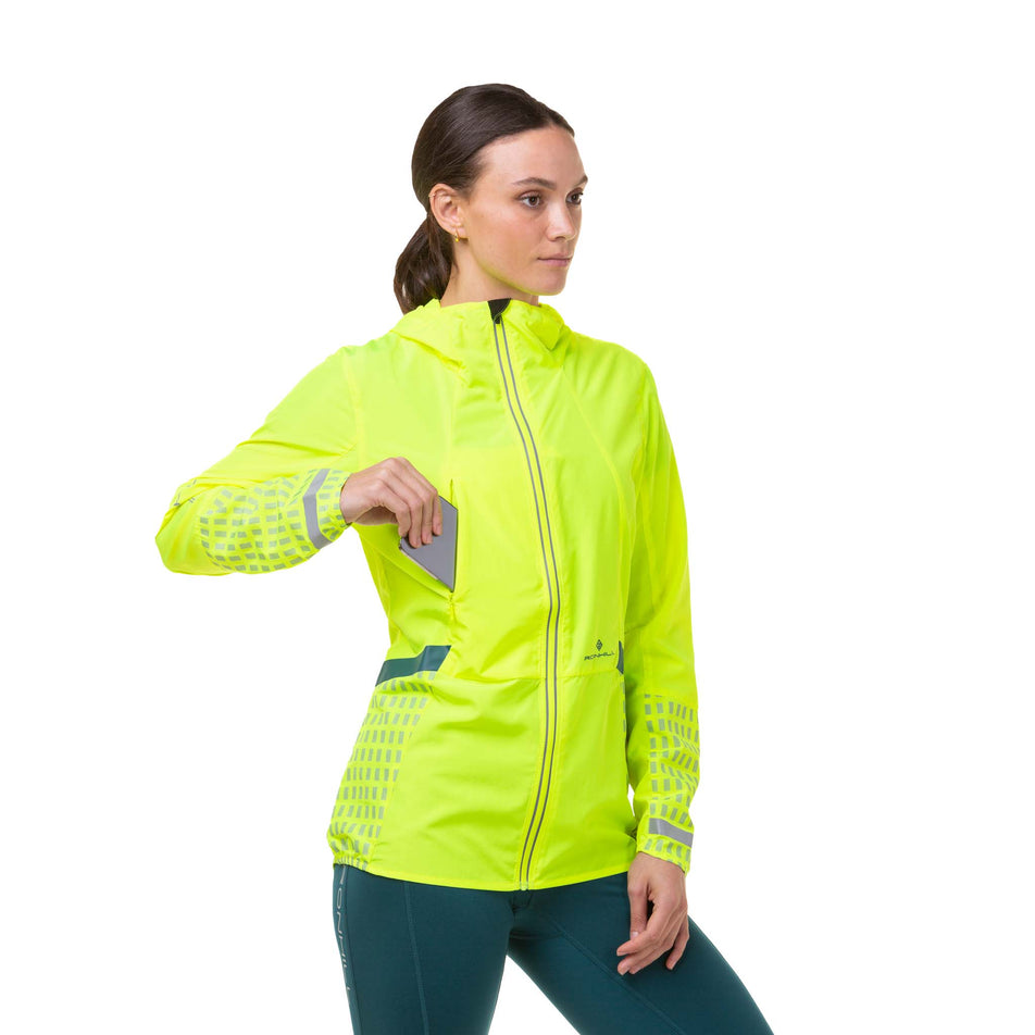 Front view of a model wearing a Ronhill Women's Tech Afterhours Jacket in the Fluo Yellow/Deep Lagoon/Reflect colourway. Demonstrating that a phone can be stored in the pocket on the front-right of the jacket. (8019130286242)
