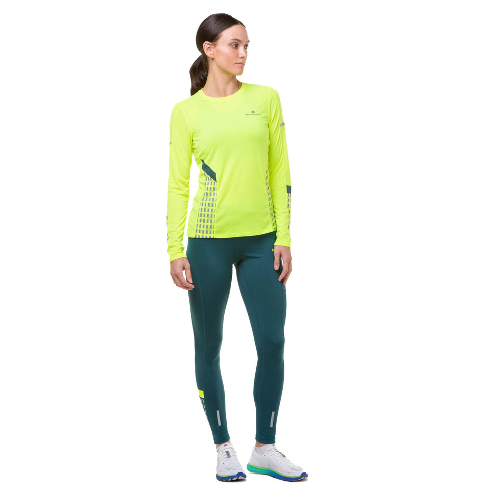 Front view of a model wearing a Ronhill Women's Tech Afterhours L/S Tee  in the Fluo Yellow/Deep Lagoon/Reflect colourway. The model is also wearing a pair of green tights and white running shoes. (8023196041378)