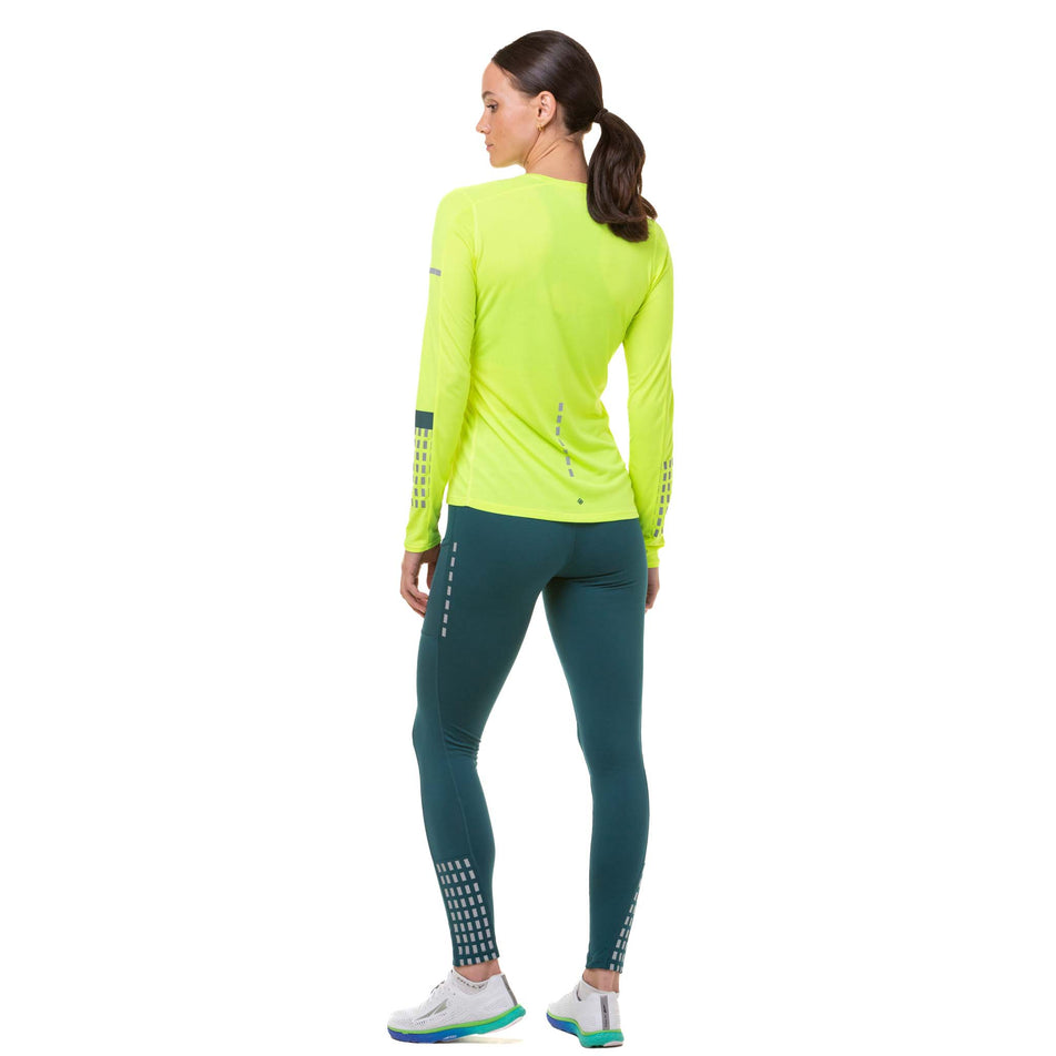 Back view of a model wearing a Ronhill Women's Tech Afterhours L/S Tee in the Fluo Yellow/Deep Lagoon/Reflect colourway. The model is also wearing a pair of green tights and white running shoes. (8023196041378)