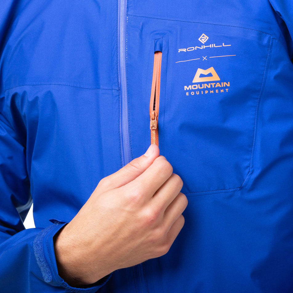 Close-up view of a model demonstrating that there is a zipped chest pocket on a Ronhill Men's Tech Gore-Tex Mercurial Jacket in the Cobalt/Copper colourway. (8032222019746)