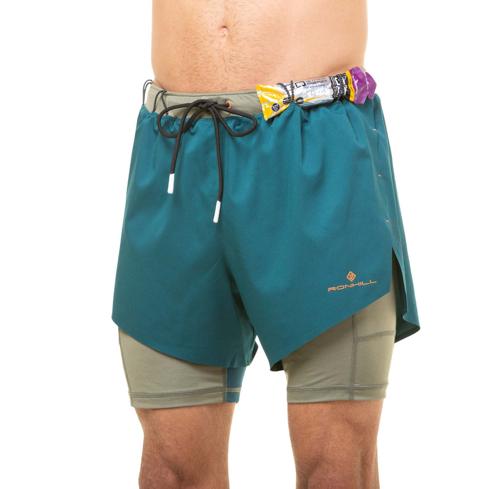 Front view of a model wearing a pair of Ronhill Men's Tech Distance Twin Shorts in the Deep Lagoon/Woodland colourway. There is an energy gel stored in the elasticated loops on the waistband. (8048123084962)