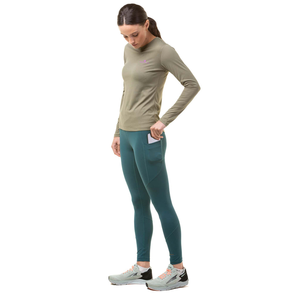 Side view of a model demonstrating that a phone can be stored in the left-thigh stretch pocket on a pair of Ronhill Women's Tech Tights in the Deep Lagoon/Copper colourway. Model also wearing a green Ronhill long sleeve top and grey Altra running shoes. (8024370315426)