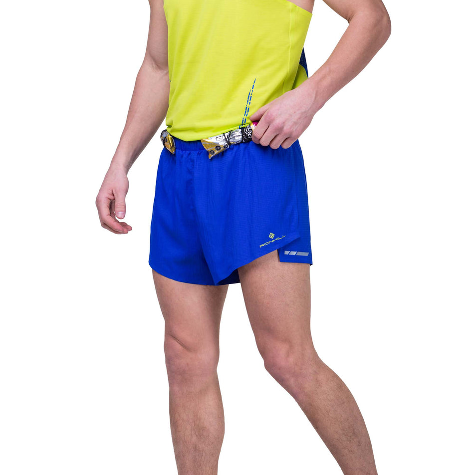 Front view of a model wearing the Ronhill Men's Tech Race Short in the Azurite/Citrus colourway, and demonstrating that energy gels can be stored in the elasticated front loops. Model is also wearing a Ronhill running vest. (8159260410018)