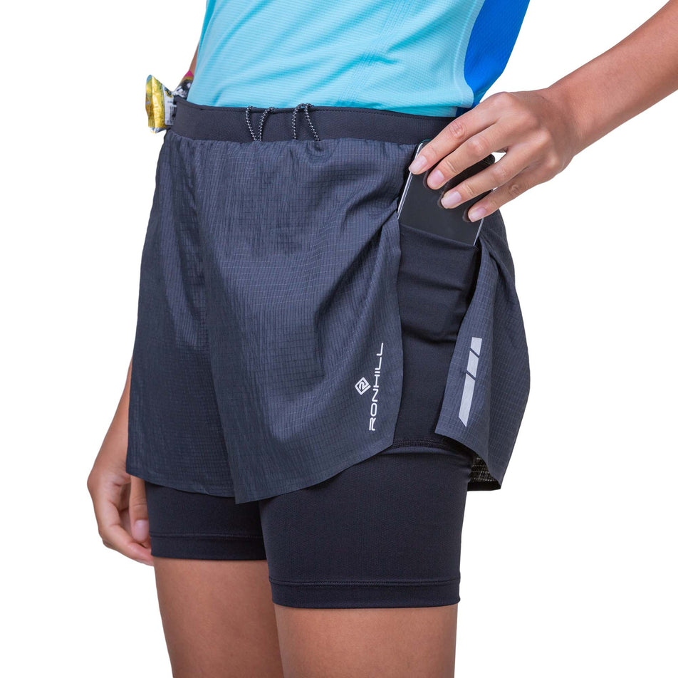 Front view of a model wearing the Ronhill Women's Tech Race Twin Short in the All Black colourway, and demonstrating that a phone can be stored in the stretch pocket on the left leg. Model is also wearing a running top. (8158813323426)