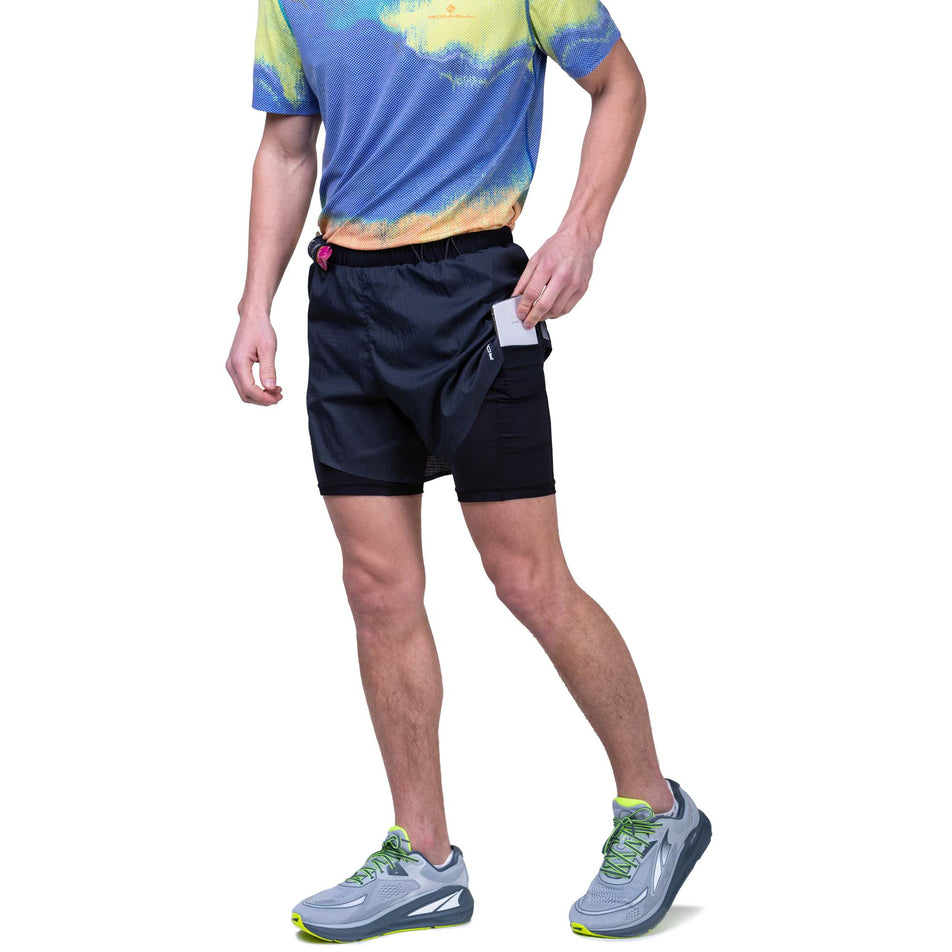 Front view of a model wearing the Ronhill Men's Tech Race Twin Short in the All Black colourway. Model demonstrating that a phone can be stored in the stretch pocket on the left side of the inner layer. Model also wearing a Ronhill top and Altra running shoes.  (8159263883426)