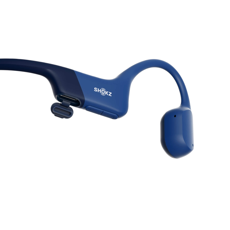 Close-up of the right earbud from a pair of Shokz Unisex OpenRun Wireless Bone Conduction Headphones in the Blue clourway. Image shows a USB-Type C charging port. (8541454467234)