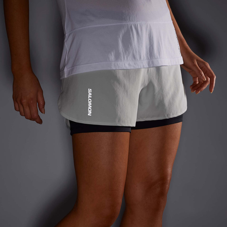 Front view of a model wearing a pair of Salomon Women's Cross 2in1 Shorts in the Oyster Mushroom colourway. Photo taken in low light so that the reflective Salomon logo can be seen.  (7892130201762)