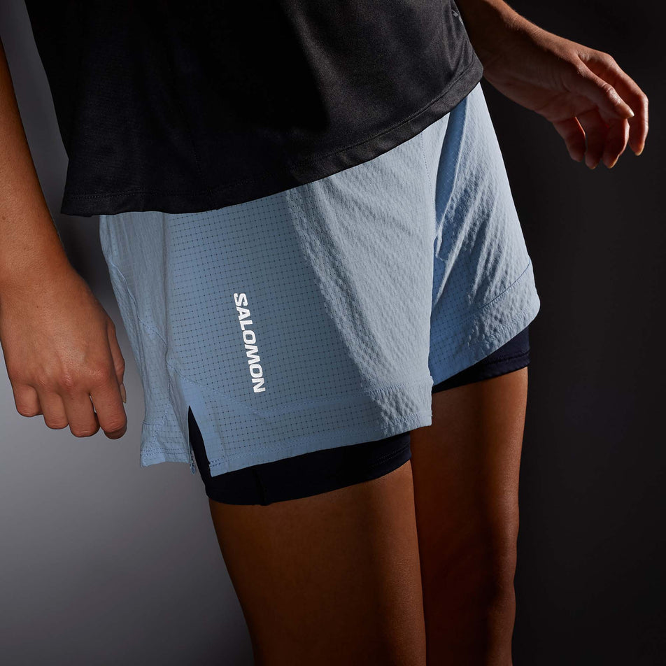 Close-up front view of a model wearing a pair of Salomon Women's Sense Aero 2in1 Shorts in low light conditions - to show the reflective Salomon logo. Chambray Blue/Deep Black colourway  (7892306952354)