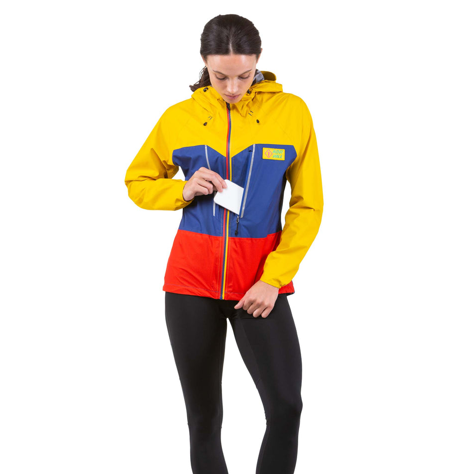 Front view of a model wearing and taking a phone out of the chest pocket on a Ronhill Women's Tech Fortify Jacket in the Dark Cobalt/Solar colourway (7746201976994)